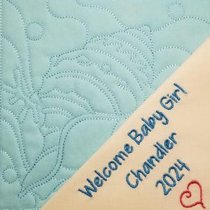 May include: A light blue quilted fabric with a floral design. A cream-colored fabric with the text "Welcome Baby Girl Chandler 2024" and a red heart is sewn on top of the blue fabric.