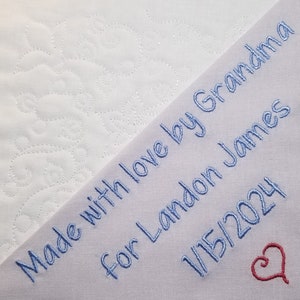 May include: A white fabric with a blue embroidered message that reads "Made with love by Grandma for Landon James 1/15/2024" and a red heart.