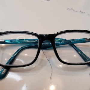 Könnte beinhalten: Rechteckige Brille mit glänzend schwarzer Front und durchscheinenden türkisfarbenen Bügeln. Auf der Innenseite des Bügels ist der Text XOXO, CE und Turquoise (BLTQ) 54-16-140 aufgedruckt. Die klaren Gläser sind in den schwarzen Kunststoffrahmen gefasst.