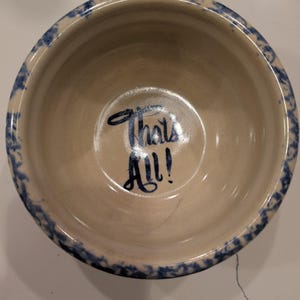 May include: A round ceramic bowl with a cream-colored interior and a blue sponge-painted rim. The center of the bowl features the dark blue text "That's All!" written in a cursive script. The bowl has a smooth, glazed finish throughout.