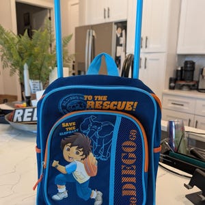 May include: A dark blue rolling backpack with a light blue extendable handle and wheels. The front features a graphic of Diego, an elephant outline, and the text "Go, Diego, Go! To The Rescue!" and "Save The Elephant" with orange accents.