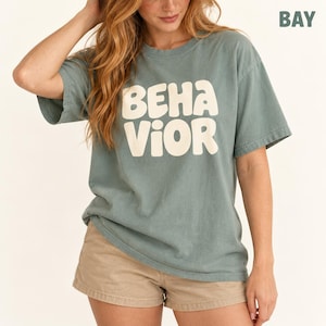 Op de afbeelding: Een stoffig blauw t-shirt met korte mouwen en het woord BEHAVIOR in grote, crèmekleurige, afgeronde blokletters op de borst. Het t-shirt heeft een ronde hals en een relaxte pasvorm, gecombineerd met een beige korte broek.