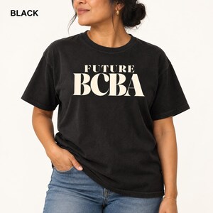 Könnte beinhalten: Ein schwarzes Kurzarm-T-Shirt mit dem Text FUTURE BCBA, der in einer großen, fetten, cremefarbenen Serifenschrift auf der Brust gedruckt ist. Das Kleidungsstück hat einen klassischen Rundhalsausschnitt und eine lockere Passform, kombiniert mit einer blauen Jeans.