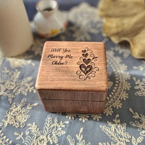 May include: A wooden ring box with the text "Will You Marry Me Chloe?" engraved on the lid. The box features a decorative heart design. The box is made of light brown wood.