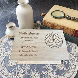May include: A light-colored wooden baptism plaque with engraved text and a religious symbol. The plaque includes the name "Stella Yasmine", the date "August 31, 2025", and the church name. Decorative elements and a vintage aesthetic.
