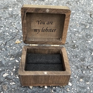 Pu&ograve; includere: Una scatola per anelli in legno con un interno in velluto nero. Il coperchio &egrave; aperto e l'interno &egrave; inciso con le parole "You are my lobster".