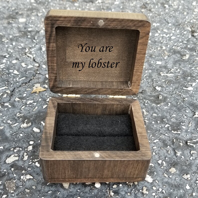 Pu&ograve; includere: Una scatola per anelli in legno con un interno in velluto nero. Il coperchio &egrave; aperto e l'interno &egrave; inciso con le parole "You are my lobster".