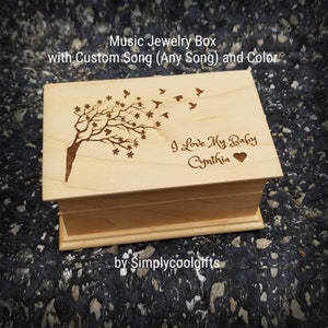 May include: A light-colored wooden music jewelry box with a tree and birds design. The text "I Love My Baby Cynthia" is engraved on the lid. The box is labeled "Music Jewelry Box with Custom Song (Any Song) and Color" and "by Simplycoolgifts".