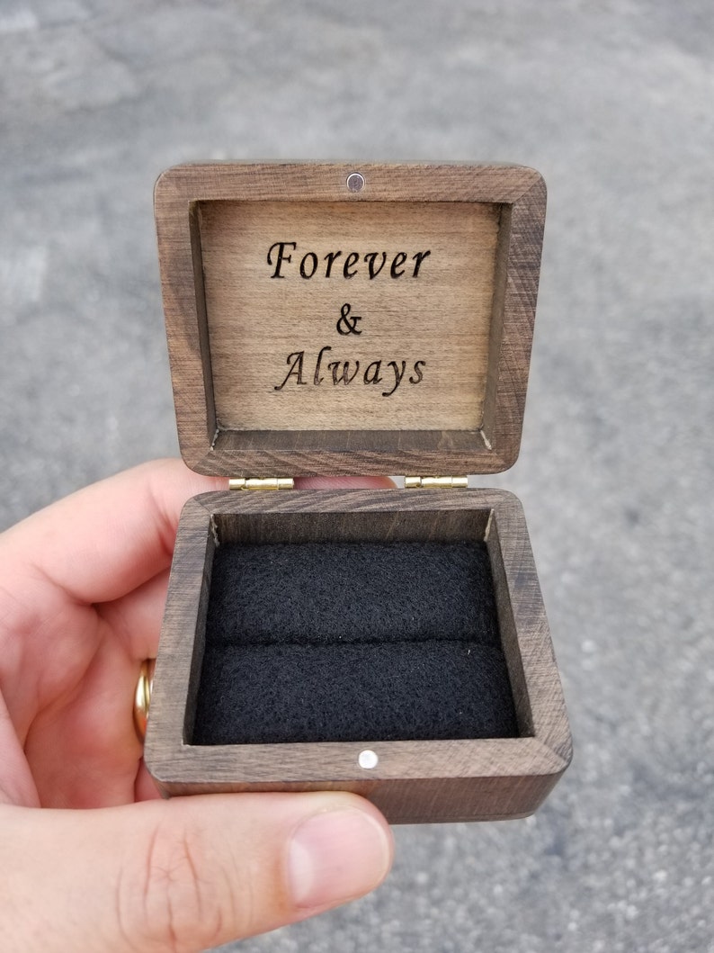 Pu&ograve; includere: Una scatola per anelli in legno con un interno in velluto nero. Il coperchio &egrave; aperto e l'iscrizione "Forever & Always" &egrave; visibile.