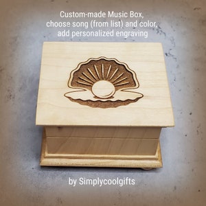 May include: A wooden music box with a pearl in a seashell design on the lid. The box is made to order and can be personalized with a song and engraving. The text on the box reads "Custom-made Music Box, choose song (from list) and color, add personalized engraving by Simplycoolgifts".