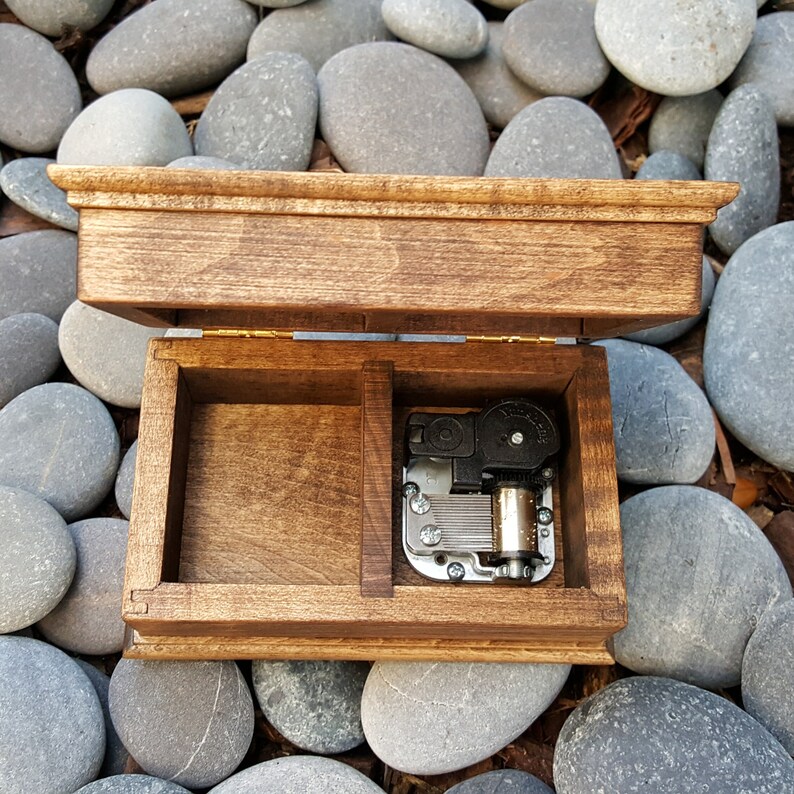 Puede incluir: Una caja de m&uacute;sica de madera con una tapa con bisagras, abierta para revelar un mecanismo musical plateado. La caja es de madera marr&oacute;n oscura y tiene un dise&ntilde;o sencillo.