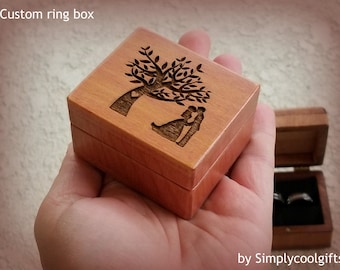 May include: A wooden ring box with a laser-etched design of a tree and a couple embracing. The box is square, with a hinged lid. The text "Custom ring box" is at the top.