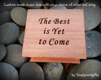 May include: A wooden music box with the inscription "The Best is Yet to Come" on the top. The box is made of a light brown wood and has a hinged lid.