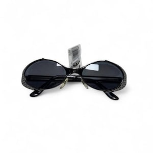 May include: Black sunglasses with oval-shaped dark lenses and a glossy black frame. The side shields feature a perforated pattern. A white product tag with a barcode and the text B-54 is attached to the bridge between the two lenses.