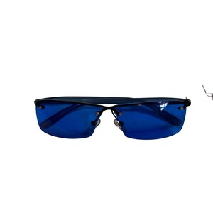 May include: Rimless sunglasses featuring vibrant blue lenses and a thin black metal frame. The design includes a straight bridge, adjustable nose pads, and slender black temples. The lenses are secured to the frame with small silver-toned screws.