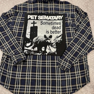 May include: A navy blue and white plaid long-sleeve button-down shirt featuring a large rectangular black patch on the back. The patch displays a graphic of a cat and a tombstone with the text PET SEMATARY Sometimes dead is better.