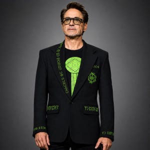 May include: A black blazer featuring bright green runic-style embroidery along the lapel, cuffs, and pocket flaps, paired with a black t-shirt displaying a large, geometric green graphic. The blazer has a single-button closure and notched lapels.