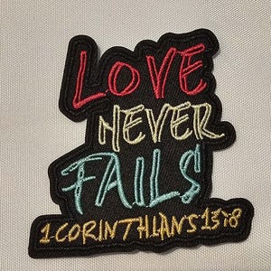 Op de afbeelding: Een zwarte geborduurde patch met de tekst LOVE in rood, NEVER in geel en FAILS in lichtblauw, met de vermelding 1 CORINTHIANS 13:8 in het geel onderaan. De patch heeft een zwarte rand en een basis van gestructureerde stof.