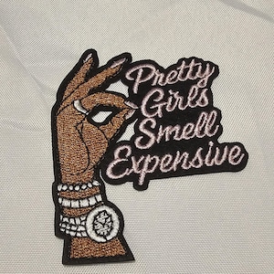May include: An embroidered patch featuring a brown-toned hand making an okay gesture, adorned with white bracelets and a watch. The text Pretty Girls Smell Expensive is stitched in light pink cursive lettering against a black background.