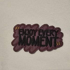 May include: An embroidered patch featuring a dark purple, cloud-shaped background with a textured, scribbled appearance. The black, bold, sans-serif text reads "ENJOY EVERY MOMENT" across the center, accented by two small, gold-colored butterfly icons.