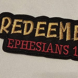 May include: A black embroidered patch with a gold-colored stitched border and the word REDEEMED in gold thread. Below the word, the text EPHESIANS 1-7 is stitched in red thread. The patch has a textured fabric base and a die-cut shape.