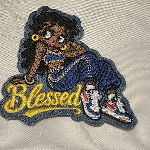 Puede incluir: Un parche bordado que muestra un personaje de dibujos animados de piel oscura y cabello negro, con un top corto azul y pantalones azules, sentado sobre el texto bordado en amarillo Blessed. El personaje lleva aros dorados y zapatillas.