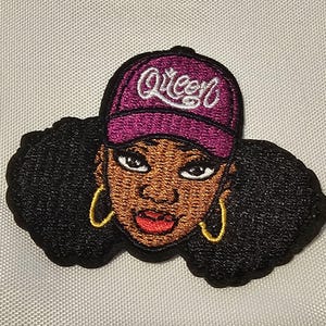 Peut inclure: Un écusson brodé représentant une personne à la peau foncée, aux cheveux afro noirs volumineux et portant des créoles dorées. La personne porte une casquette de baseball magenta avec le mot Queen brodé en écriture blanche sur le devant.