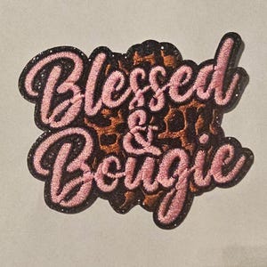May include: An embroidered patch featuring the text Blessed & Bougie in a light pink script font. The lettering is outlined in black and set against a brown and black leopard print background with a dark, glittery border surrounding the entire shape.
