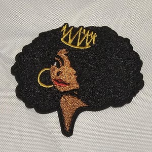 May include: An embroidered patch featuring a profile view of a person with dark skin and voluminous black hair. The figure wears a gold crown on top of the hair and a gold hoop earring. The lips are highlighted in red, and the entire design is outlined in black.