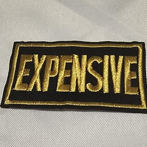 Puede incluir: Un parche rectangular bordado con fondo negro y la palabra EXPENSIVE cosida en hilo dorado metálico. El parche está bordeado por un marco grueso bordado en oro, creando un contraste marcado entre la tela oscura y las letras en relieve.