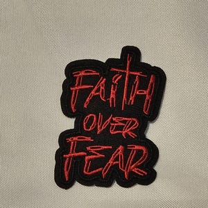 May include: A black embroidered patch featuring the red stitched text FAITH OVER FEAR. The word FAITH is positioned at the top with a cross integrated into the letter T, followed by the words OVER and FEAR stacked vertically below it.