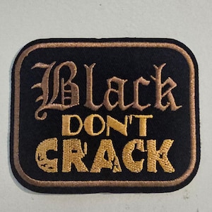 Peut inclure: Un écusson brodé rectangulaire aux coins arrondis avec un fond noir et une bordure dorée. Le texte "Black DON'T CRACK" est brodé en fil doré, avec "Black" dans une police gothique décorative et "DON'T CRACK" en lettres capitales droites.