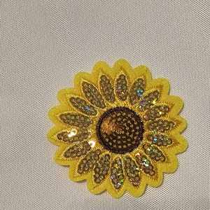 May include: A sunflower-shaped embroidered patch featuring bright yellow petals and a dark brown center. The design is embellished with iridescent, light-reflecting sequins throughout the petals and the center, creating a shimmering effect.