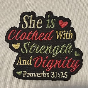 May include: A black embroidered patch featuring the text She is Clothed With Strength And Dignity Proverbs 31:25. The lettering uses a mix of gold, red, and green thread colors, accented by small embroidered heart shapes in red, gold, and green.