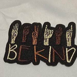 Peut inclure: Un écusson brodé noir représentant six mains de différentes carnations effectuant des gestes en langue des signes au-dessus du texte BE KIND, brodé en fil beige clair sur un fond en tissu noir.