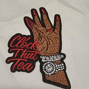 Puede incluir: Un parche bordado que muestra una mano de tono marrón con uñas rojas, luciendo un reloj plateado y una pulsera de cuentas. El texto "Clock That Tea" está bordado en letras cursivas rojas a la izquierda del diseño de la mano.