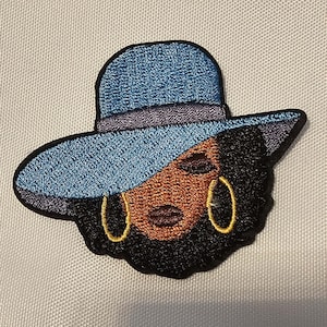 Puede incluir: Un parche bordado que muestra a una persona de piel oscura y cabello negro con un sombrero azul de ala ancha y una banda oscura. La figura tiene los ojos cerrados, labios oscuros y grandes aros dorados, todo delineado con hilo negro sobre fondo blanco.