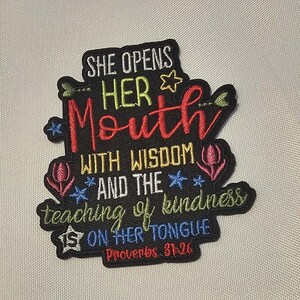 May include: A black embroidered patch featuring the text SHE OPENS HER MOUTH WITH WISDOM AND THE teaching of kindness IS ON HER TONGUE Proverbs 31:26 in red, yellow, green, and blue thread, accented with small embroidered stars and flower icons.