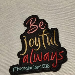 May include: A black embroidered patch featuring the text Be joyful always in a mix of pink, gold, and red thread. Below the main text, the word Thessalonians 5:16 is stitched in light blue thread. The patch has a black border and a textured finish.