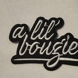 May include: A black embroidered patch featuring the white cursive text a lil' bougie. The design consists of white thread lettering stitched onto a black fabric background with a thick black border following the shape of the script text.