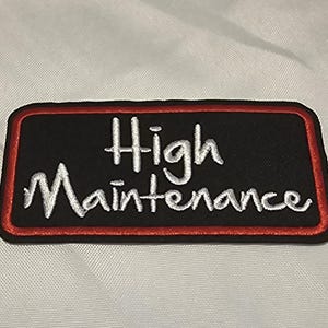 May include: A rectangular embroidered patch with a black background and a red border. The text "High Maintenance" is stitched in white thread in a cursive script font across the center of the patch. The patch has a clean, finished edge.