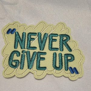 May include: An embroidered patch featuring the text "NEVER GIVE UP" in dark teal thread. The letters are surrounded by a light yellow, cloud-like stitched background with small blue quotation marks at the top left and bottom right corners.