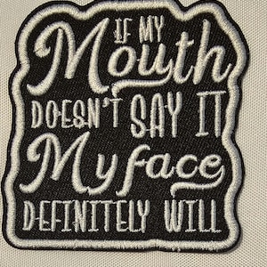 May include: A black embroidered patch with a white border and white stitched text that reads: IF MY Mouth DOESN'T SAY IT My face DEFINITELY WILL. The patch has a wavy, irregular shape and features a combination of cursive and block lettering styles.