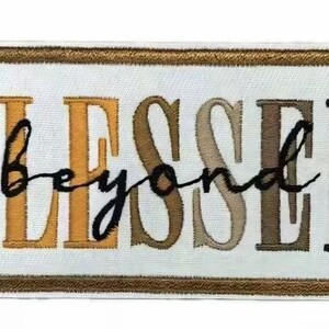 May include: A rectangular embroidered patch with a white background and a gold border. The word BLESSED is stitched in large, block letters that transition from gold to brown. The word beyond is stitched in black cursive script across the center.