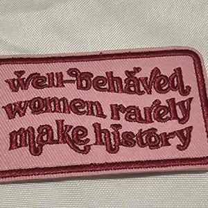 May include: A rectangular embroidered patch with a light pink background and a dark red border. The text "Well-behaved women rarely make history" is stitched in dark red thread across the centre in a stylised, decorative font.
