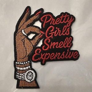 Peut inclure: Un écusson brodé représentant une main brune faisant un geste de signe ok avec des ongles rouges, une bague argentée et des bracelets argentés. Le texte Pretty Girls Smell Expensive est brodé en fil rouge sur un fond noir à côté de la main.