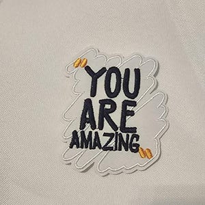May include: An embroidered patch featuring the black text YOU ARE AMAZING on a white background with a scribbled border. The design includes two small gold quotation marks at the top left and bottom right corners of the white patch area.