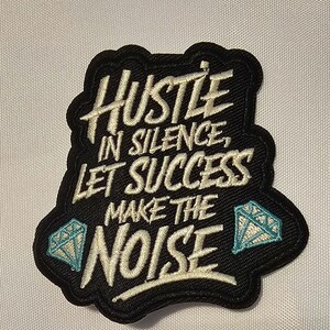 Op de afbeelding: Een zwarte geborduurde patch met de witte tekst Hustle in silence, let success make the noise. Het ontwerp bevat twee lichtblauwe diamanticonen, één linksonder en één rechtsonder, allemaal op een zwarte achtergrond.