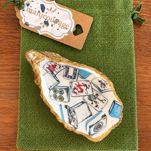 May include: A decorative oyster shell dish with a gold-painted rim and a white interior featuring a pattern of colorful mahjong tiles. The shell rests on a green burlap drawstring pouch with a brown paper tag labeled with the text mahjcoupage.