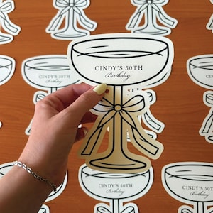 Puede incluir: Un recorte de papel troquelado con forma de copa de champán con un lazo decorativo en el tallo. La copa presenta el texto impreso CINDY'S 50TH Birthday en tinta negra. El diseño está realizado en color crema con contornos negros sobre papel.